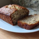 A golden brown loaf of Incredibly Moist Banana Bread, partially sliced, sitting on a white plate.