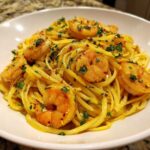 A close-up of a white bowl filled with Lemon Butter Shrimp Pasta, topped with sautéed shrimp and fresh parsley.