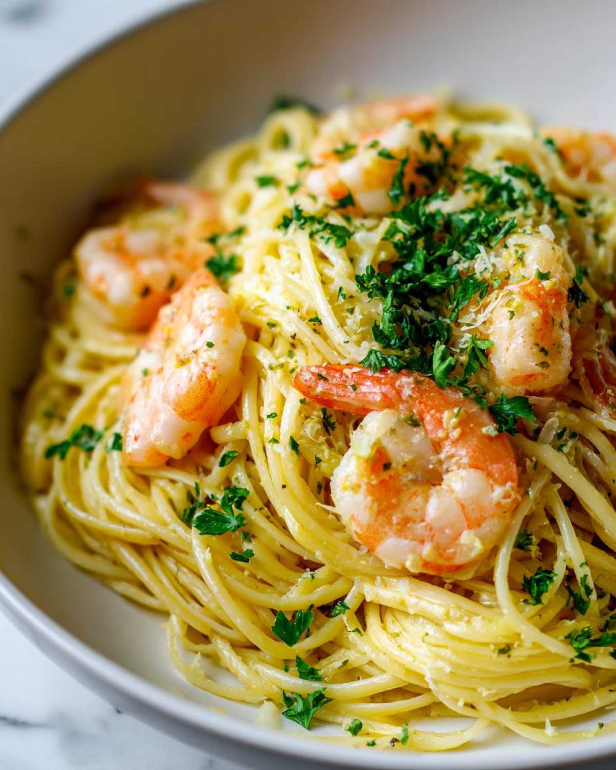 A close-up of a white bowl filled with Lemon Butter Shrimp Pasta, topped with pink shrimp and fresh parsley.