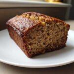 A close-up of a thick slice of moist Banana Bread with a dark, caramelized crust, served on a white plate.
