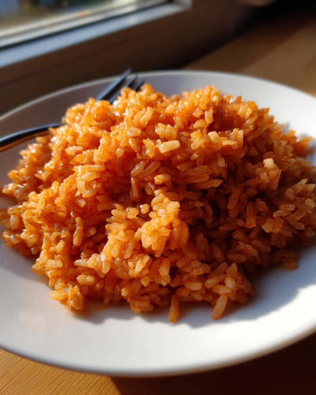 A close-up of fluffy, orange-colored Spanish rice easy recipe served on a white plate, illuminated by sunlight.