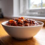 A steaming white bowl filled with rich, dark Vegan Chili Recipe, featuring visible chunks of sweet potato and beans.