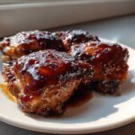 Four pieces of Sticky Honey Soy Chicken Thighs with Garlic, coated in a thick, glossy, dark brown glaze, served on a white plate.
