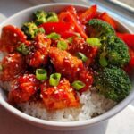 Close-up of a Sticky Sweet Chili Chicken Bowl with rice, bright red sauce, broccoli, and sesame seeds.