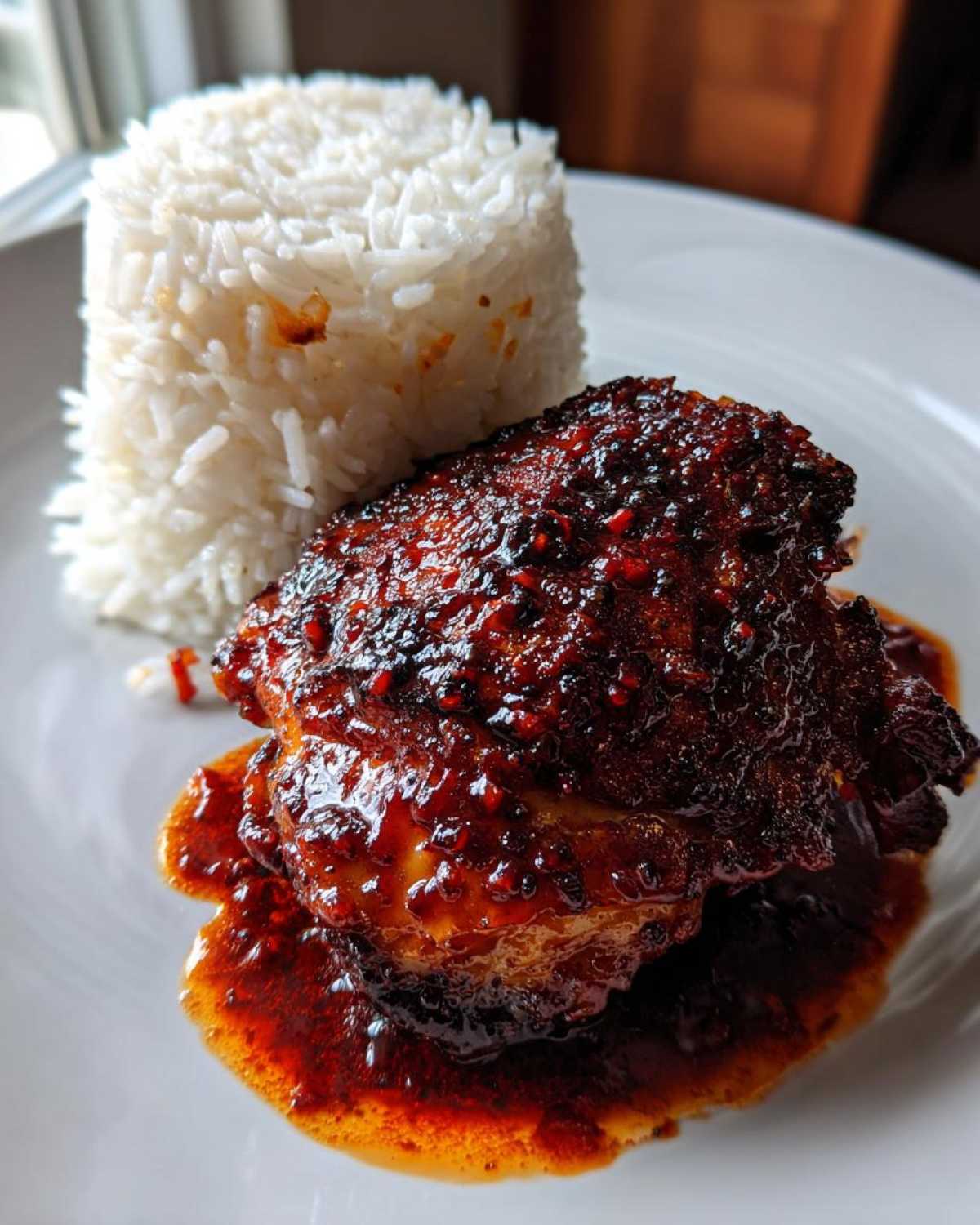 A close-up of Sticky Sweet Chili Chicken Thighs glazed with a thick, dark red sauce, served next to a molded portion of white rice.
