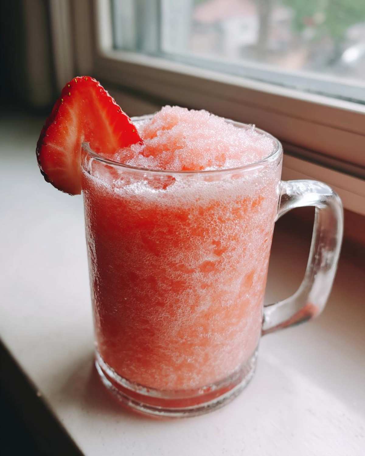 A close-up of a frosty glass mug filled with bright pink, blended Irresistible Strawberry Pina Colada Punch, garnished with a fresh strawberry slice.