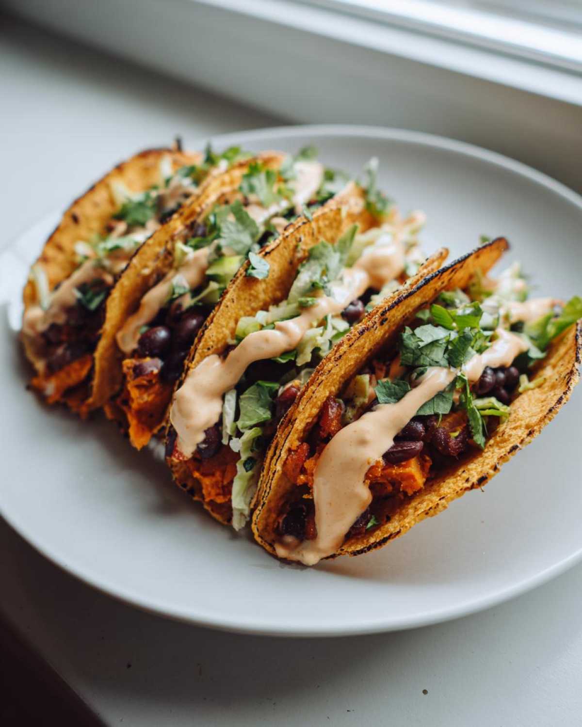 Four crispy tacos filled with sweet potato and black bean mixture, topped with slaw, cilantro, and a creamy sauce, showcasing Sweet Potato and Black Bean Tacos.