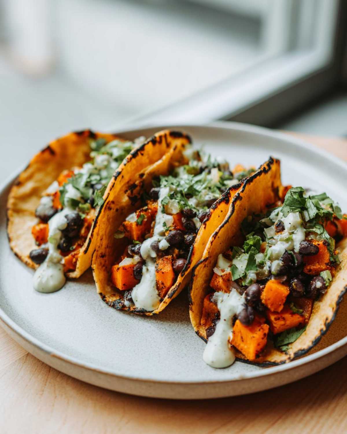 Three delicious Sweet Potato and Black Bean Tacos served on a light gray plate, topped with crema and cilantro.