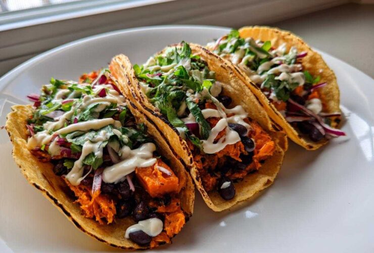 Three delicious Sweet Potato and Black Bean Tacos filled with orange sweet potato, black beans, red onion, greens, and drizzled with white sauce.