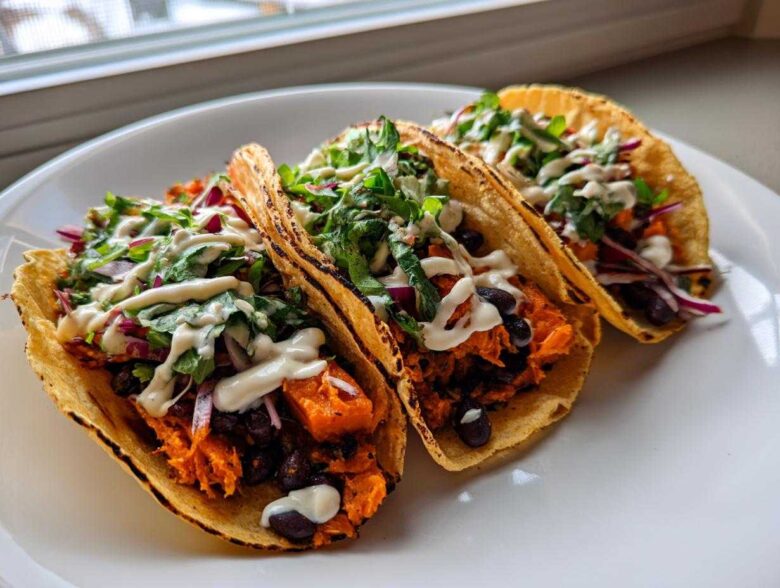 Three delicious Sweet Potato and Black Bean Tacos filled with orange sweet potato, black beans, red onion, greens, and drizzled with white sauce.