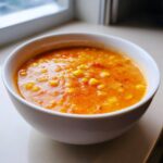 A close-up of a white bowl filled with bright orange Vegan Red Lentil Corn Chowder, showing visible corn kernels.