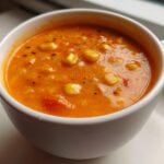 A close-up view of a white mug filled with creamy, orange Vegan Red Lentil Corn Chowder, topped with corn kernels and spices.