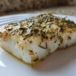 Close-up of a flaky, white Air Fryer Cod Fillets seasoned generously with dried rosemary and spices on a white plate.