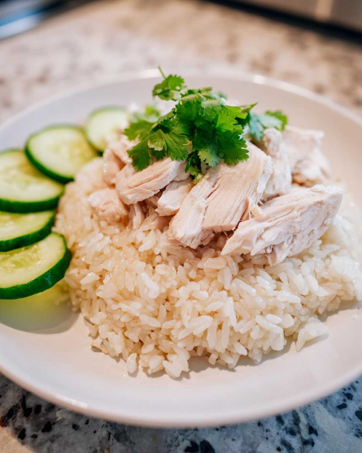 A plate of delicious Chicken Rice featuring sliced poached chicken over fragrant rice, garnished with cilantro and cucumber slices.