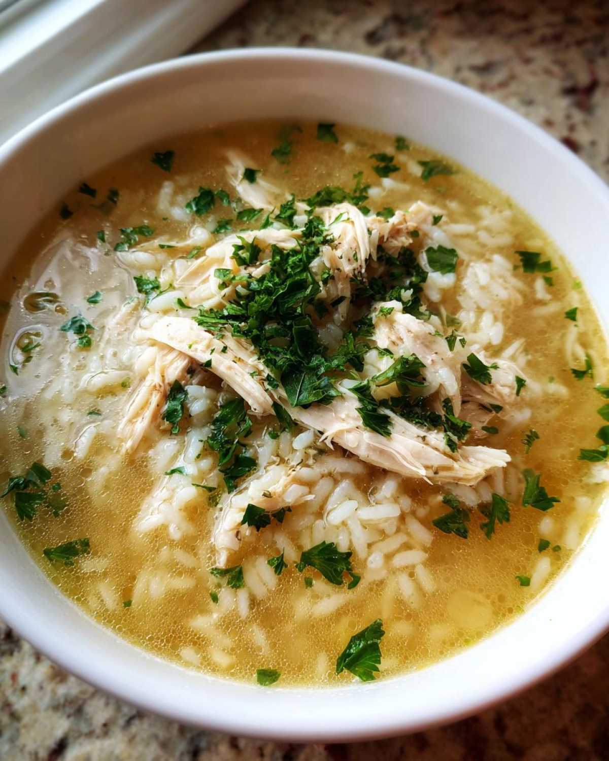 Close-up of a white bowl filled with flavorful Chicken Rice Soup, topped with shredded chicken and fresh parsley.