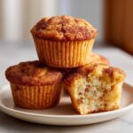 A stack of Apple Cinnamon Greek Yogurt Muffins topped with cinnamon sugar, one muffin is bitten into showing the soft interior.