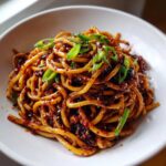 A close-up of thick, glossy Asian Garlic Noodles tossed in a dark, spicy sauce, topped with fresh green onions.