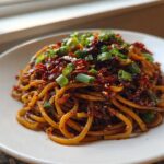 A close-up of glossy, saucy Asian Garlic Noodles topped generously with chili flakes and sliced green onions.