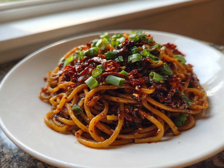 A close-up of glossy, saucy Asian Garlic Noodles topped generously with chili flakes and sliced green onions.