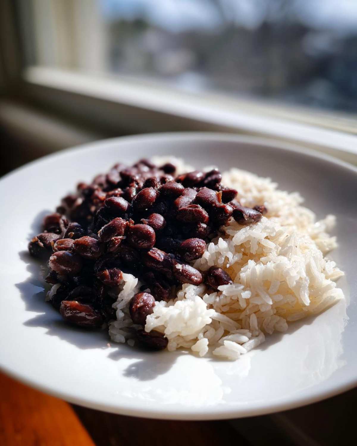 A close-up of a serving of rich, dark Black Beans and Rice on a white plate, illuminated by natural light from a window.