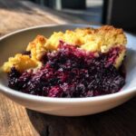 A close-up of a serving of rich Blackberry Cobbler with a golden, crumbly topping in a white bowl.