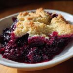 A close-up of a serving of rich, dark Blackberry Cobbler with a golden, crumbly topping in a white bowl.