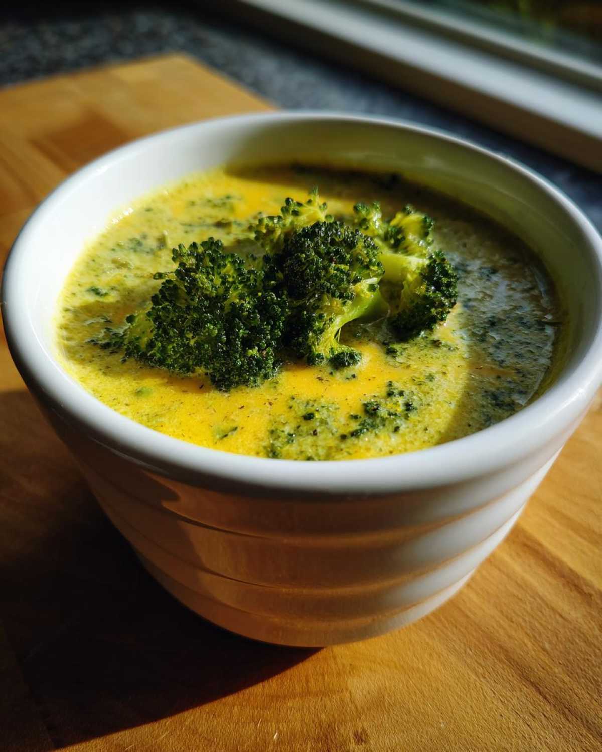 Close-up of a white bowl filled with rich, yellow Broccoli and Cheddar Soup, topped with fresh broccoli florets.