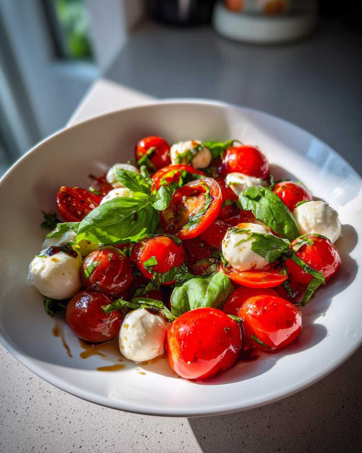 Close-up of a white bowl filled with vibrant Caprese Pasta Salad ingredients: cherry tomatoes, mozzarella balls, and fresh basil leaves drizzled with balsamic glaze.
