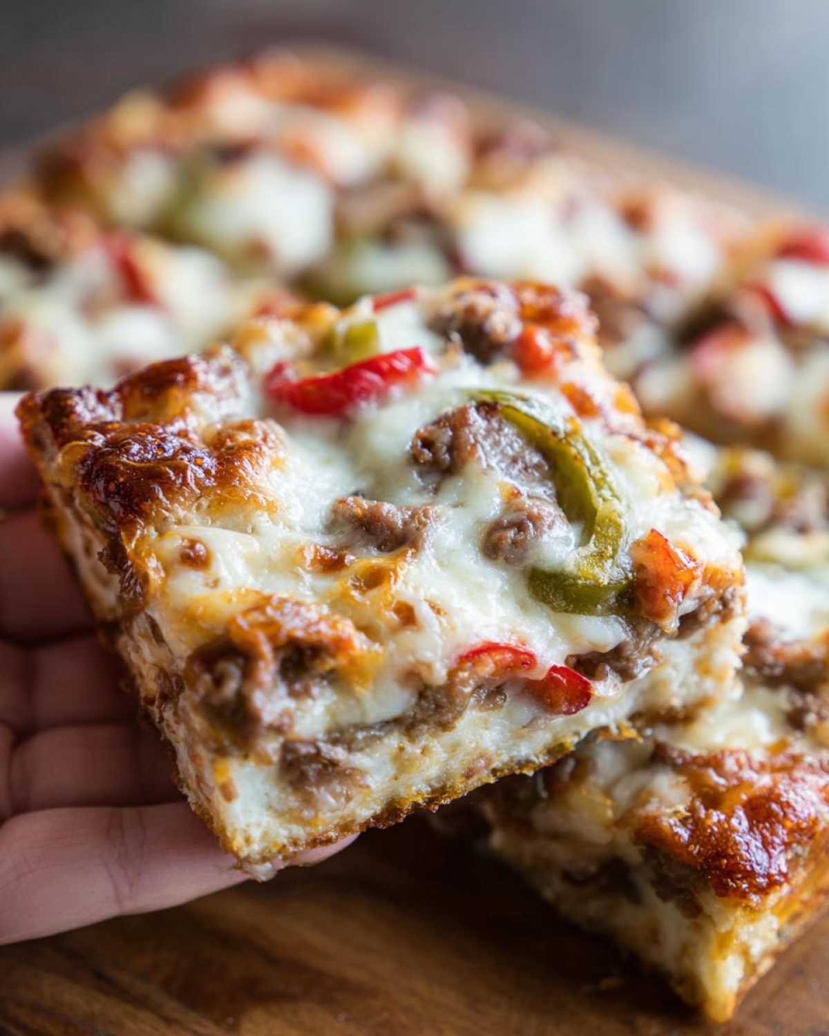 A hand holding up a thick, cheesy slice of Cheesesteak casserole topped with melted cheese and peppers.