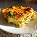 A square slice of Cheesy Veggie Quinoa Casserole topped with melted, browned cheese, served on a white plate.