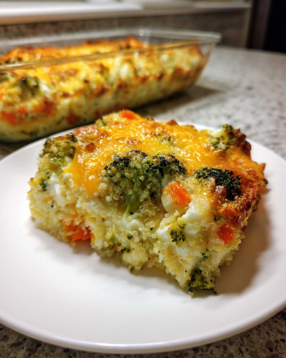 A square serving of Cheesy Veggie Quinoa Casserole topped with melted cheddar cheese, broccoli, and carrots.