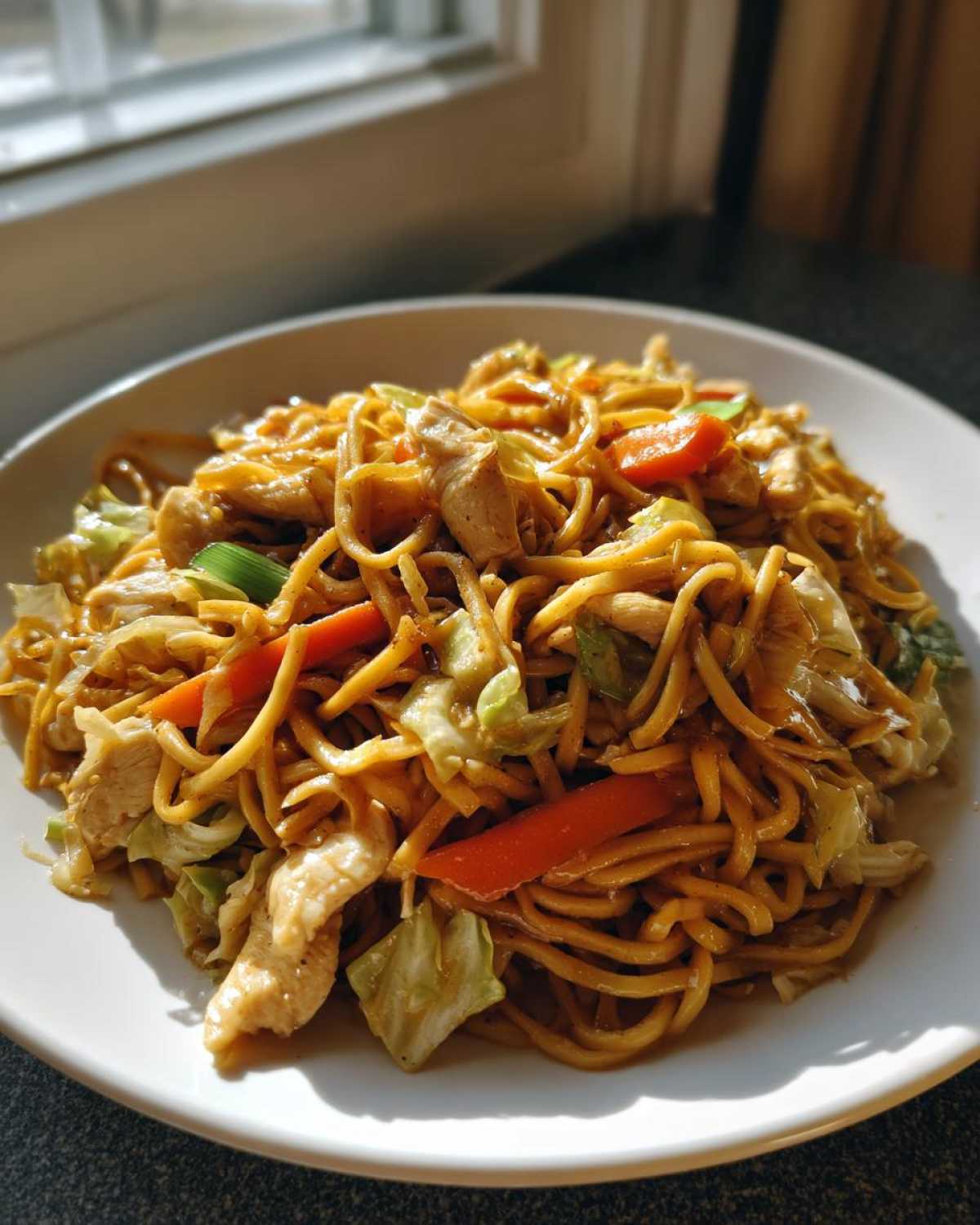 A close-up of a white plate filled with glistening Chicken Lo Mein noodles, mixed with chicken pieces, carrots, and cabbage.