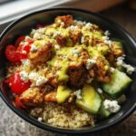 A close-up of a delicious Chicken Quinoa Bowl topped with seasoned chicken, feta, tomatoes, cucumber, and a yellow dressing.