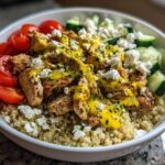 A vibrant Chicken Quinoa Bowl featuring grilled chicken pieces topped with yellow sauce and feta, served over quinoa with cherry tomatoes and cucumbers.