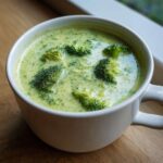 A close-up of creamy Broccoli and Cheddar Soup with visible florets in a white mug on a wooden surface.