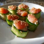 Seven pieces of Cucumber Sushi, wrapped in thin cucumber strips and topped with smoked salmon and sesame seeds, arranged on a white plate.