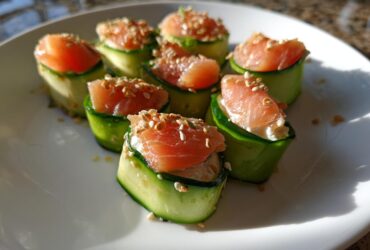 Seven pieces of Cucumber Sushi, wrapped in thin cucumber strips and topped with smoked salmon and sesame seeds, arranged on a white plate.