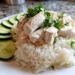 A plate of moist, sliced Chicken Rice topped with fresh parsley and served with sliced cucumbers on the side.