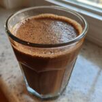 Close-up of a freshly blended, frothy Coffee Smoothie in a clear glass resting near a window.