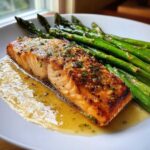 Close-up of a perfectly cooked Garlic Butter Baked Salmon Dinner fillet topped with herbs, resting in butter sauce next to bright green asparagus.