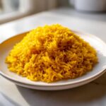 A close-up of a perfectly cooked mound of bright yellow Garlic Turmeric Rice served on a light-colored plate.