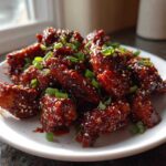 A plate piled high with sticky, glazed Korean BBQ Chicken pieces, garnished with sesame seeds and green onions.