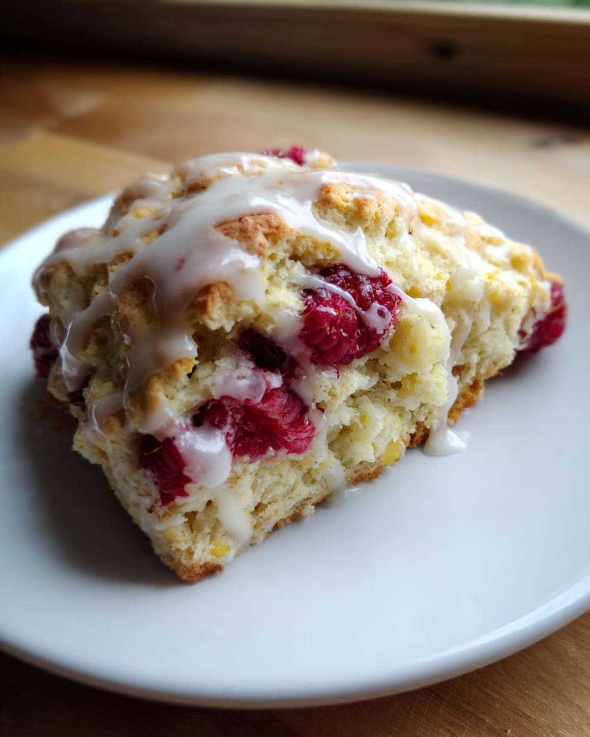 A close-up of a moist Lemon Raspberry Scone topped with a thick white glaze, served on a white plate.