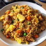 Close-up of a white bowl filled with savory Chicken Fried Rice, featuring rice, scrambled egg, peas, and carrots.