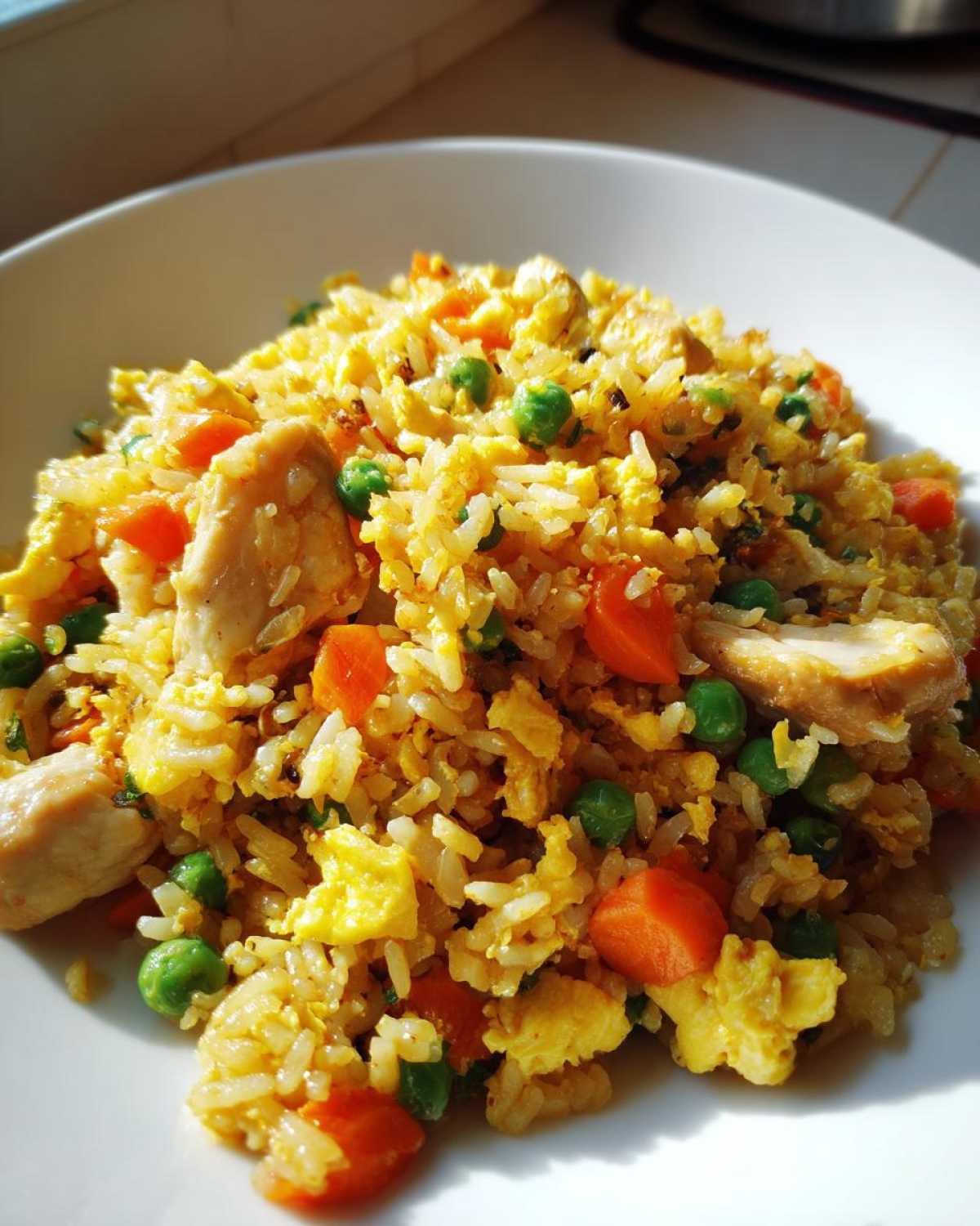 A close-up, sunlit serving of homemade Chicken Fried Rice featuring yellow rice, chunks of white chicken, bright orange carrots, and green peas.