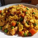 A close-up of vibrant, yellow-seasoned Chicken Fried Rice featuring chunks of chicken, bright green peas, and diced carrots on a white plate.