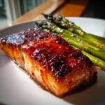 Close-up of a perfectly cooked fillet of Gochujang Honey Glazed Salmon topped with a thick, shiny red sauce, served next to roasted asparagus.