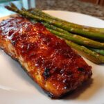 Close-up of a glistening Gochujang Honey Glazed Salmon fillet served with roasted asparagus on a white plate.