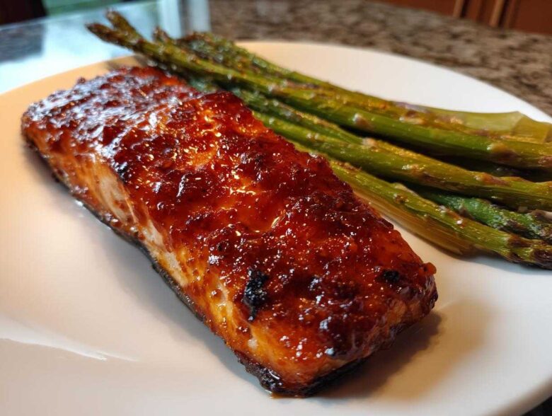 Close-up of a glistening Gochujang Honey Glazed Salmon fillet served with roasted asparagus on a white plate.
