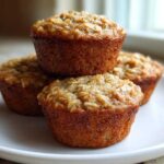 A stack of four golden-brown Healthy Banana Oatmeal Muffins topped with visible oats, served on a white plate.
